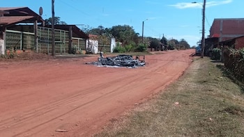 Restos calcinados de un vehículo en el centro de una calle de tierra. Se observan casas, vegetación, postes y un camión blanco al fondo a la derecha