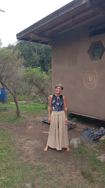 Mujer sonriente de cabello gris corto, top floral y pantalones anchos, descalza, posa frente a casa rústica con pared de tierra y diseño en espiral. Entorno rural