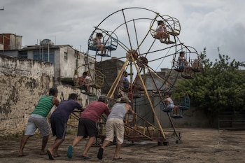 Cuatro hombres empujan la estructura de una rueda de la fortuna de metal mientras varios niños se encuentran sentados en las canastas.