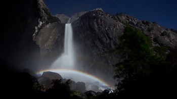 Cascada de agua blanca cae por acantilado rocoso bajo un cielo nocturno con estrellas. Un arcoíris pálido es visible en la base de la cascada