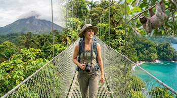 Una mujer con sombrero, mochila y cámara camina sonriente por un puente colgante en la jungla; al fondo, el volcán Arenal, un perezoso y una playa tropical.