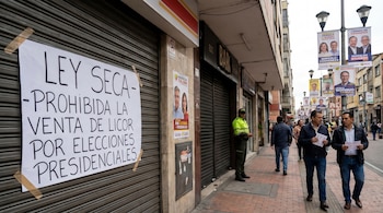 Un cartel blanco en un local cerrado anuncia la "Ley Seca" por elecciones presidenciales en Colombia, con personas y un policía en la calle, y pancartas políticas.