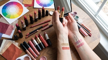 Una persona sostiene un espejo y un labial sobre una mesa de madera con varios labiales, delineadores de labios y pigmentos. Se ven muestras de color en los antebrazos, una rueda de colores y trozos de tela.