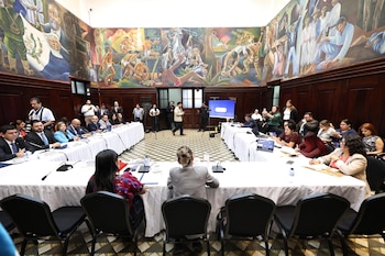 Personas sentadas alrededor de mesas con manteles blancos en un salón con micrófonos, pantallas y murales en las paredes