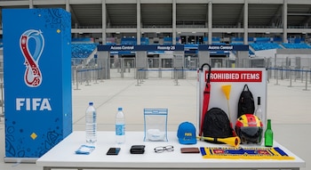 Mesa con artículos permitidos (botella vacía, teléfono, gafas) y prohibidos (paraguas, casco) frente a un puesto de control en un estadio, con una pancarta de FIFA.