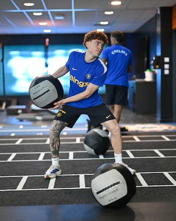 Hombre con cabello rojizo, camiseta azul del Chelsea, pantalón negro y tatuajes en la pierna, sostiene un balón medicinal en un gimnasio
