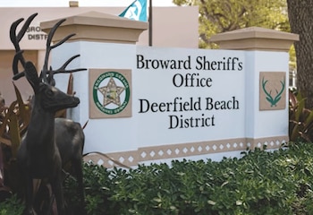 Un cartel blanco con el texto "Broward Sheriff's Office Deerfield Beach District", una estatua de ciervo y arbustos verdes