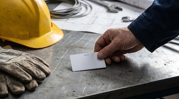 Una mano adulta con manga azul oscuro deposita una credencial de empleado blanca sobre una mesa metálica industrial. Un casco amarillo y guantes están cerca.