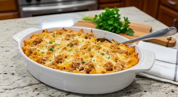 Primer plano de una cazuela ovalada blanca con pasta, atún y queso gratinado, espolvoreado con perejil, sobre una mesada de granito en una cocina.