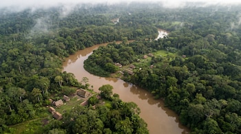 Vista aérea de un río marrón entre la selva tropical densa, con casas de techo de paja en la orilla y neblina sobre la copa de los árboles.