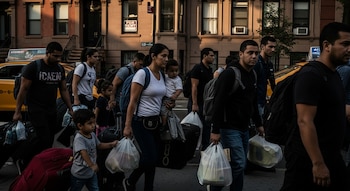 Grupo de personas, incluyendo adultos y niños, camina por una calle de la ciudad llevando equipaje y bolsas. Edificios y un taxi amarillo se ven al fondo.