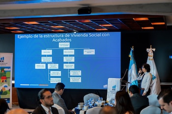 Un hombre habla con un micrófono junto a una pantalla azul con un diagrama organizacional, frente a un público sentado en mesas