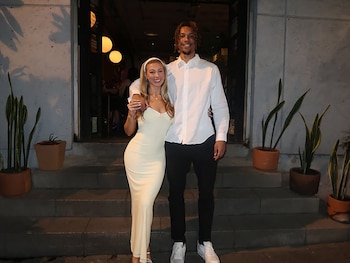 Brandon Clarke y su prometida posan de pie, sonriendo, frente a una entrada con escalones y plantas. Él lleva una camisa blanca y ella un vestido largo claro