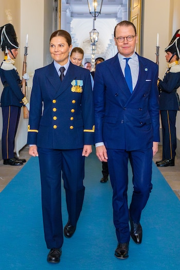 Una mujer en uniforme naval azul marino con medallas y un hombre en traje azul oscuro caminan. Dos guardias uniformados con rifles están a los lados