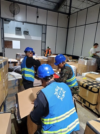 Interior de un almacén con varias personas en cascos azules y chalecos reflectantes. Inspeccionan y organizan cajas de cartón apiladas en palets