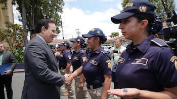 Un hombre de traje saluda a una mujer con uniforme de policía. Al fondo, otras mujeres policía en uniforme. Una oficial sostiene un objeto pequeño y redondo