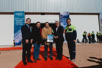 Cinco hombres con trajes y abrigos posan sobre una alfombra roja frente a un depósito industrial con carteles impresos de Posco Argentina