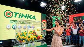Mujer con micrófono señala una tómbola con esferas de colores en un escenario con pantalla gigante que dice Tinka, público y confeti cayendo.