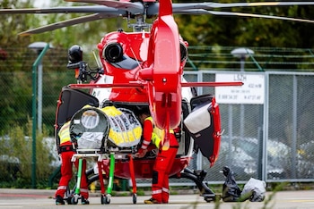 Personal médico con uniforme rojo junto a una camilla con cápsula cerrada detrás de un helicóptero rojo con las compuertas abiertas