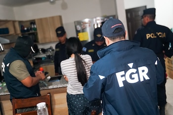 Un agente de la fiscalía con chaqueta azul y varios policías rodean a una mujer en la cocina de una vivienda