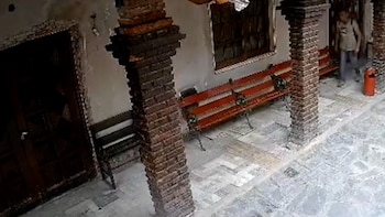 Vista de una entrada con columnas de ladrillo, un banco rojo, una puerta de madera oscura y piso de baldosas, con una persona con mochila al fondo