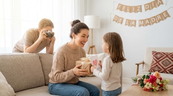 Una madre sentada en un sofá, recibiendo un regalo envuelto en papel marrón y una tarjeta con un corazón de su hija; el padre está detrás, tomando una foto.