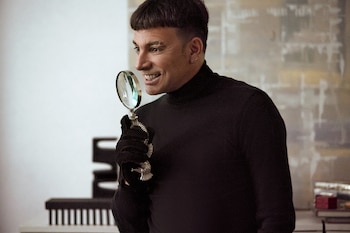 Hombre con cabello oscuro y aretes, viste suéter de cuello alto negro y guantes, sostiene lupa con mango plateado ornamentado. Fondo claro