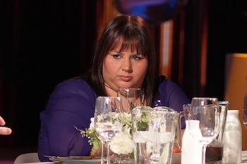 Una mujer con cabello oscuro y blusa morada permanece sentada detrás de una mesa con copas, jarras de agua y adornos florales