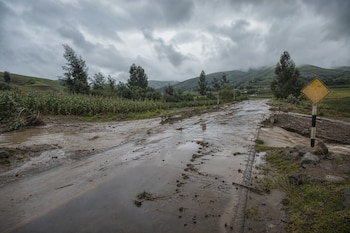 Carretera de asfalto cubierta por agua con lodo, vegetación en los costados, montañas al fondo, cielo nublado y una señal de tránsito amarilla.