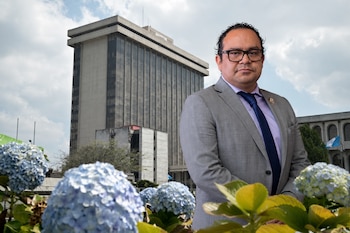 Un hombre con traje gris y anteojos posa al aire libre frente a un edificio gubernamental y plantas con flores azules.