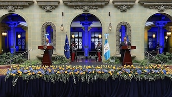 Dos hombres en podios frente a una fachada de edificio ornamentada, con banderas de la UE y Guatemala, plantas artificiales y un arreglo floral en la mesa