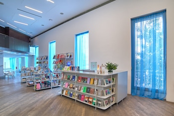 Interior de un espacio amplio y luminoso con paredes blancas y suelo de madera. Se ven varias estanterías blancas con libros y objetos a la venta, y un mostrador con flores. Grandes ventanas con cortinas azules permiten la entrada de luz