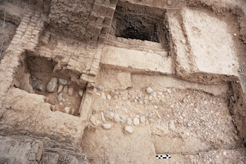 Área de excavación arqueológica con paredes de adobe, piedras, tierra expuesta, líneas de cuadrícula blancas y una regla de medición blanca y negra