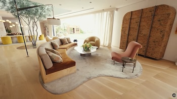 Sala de estar con sofá curvo marrón, cojines estampados, mesa de centro plateada, arreglo floral, dos sillones, armario decorado y árbol interior. Ventana al fondo