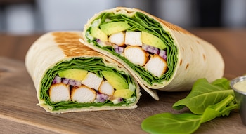 Wrap cortado en dos, con relleno de pollo, palta, hojas verdes y cebolla morada. En la superficie, hojas de espinaca y un pequeño recipiente con salsa.