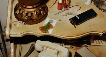 Mesa auxiliar desordenada con base de lámpara dorada, botellas, cordones, una tarjeta con un clip verde, un estuche de gafas y un teléfono de disco