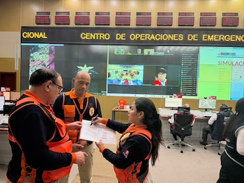 Tres personas con chalecos de Indeci revisan un documento frente a pantallas en una sala de monitoreo