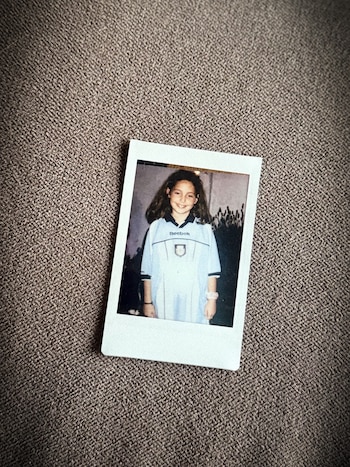 Una fotografía instantánea muestra a una niña sonriendo con cabello largo, vistiendo una camiseta deportiva azul clara con el logo de Reebok, sobre una superficie textil marrón
