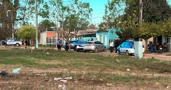 Patrulleros de policía, vehículos, personas, árboles, casas y cinta de seguridad en una calle con terreno de tierra y pasto