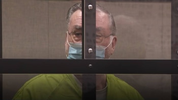 Primer plano de un hombre con gafas, mascarilla facial azul y uniforme verde, visible detrás de un panel de vidrio con un marco oscuro y tornillos