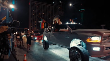 Camión pick-up blanco elevado con varias personas a bordo celebrando en una calle de noche, mientras un camarógrafo graba y la Torre de las Américas se ve al fondo