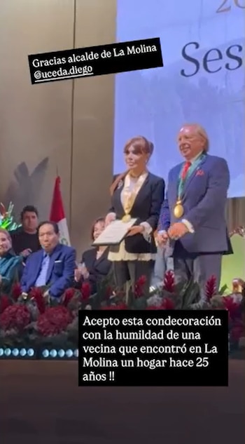 Magaly Medina y Diego Uceda en un escenario. Medina sostiene un documento y ambos llevan medallas. Hay personas sentadas en el público y una bandera