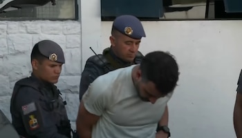 Dos agentes de policía uniformados trasladan a un hombre vestido con camiseta blanca que lleva las manos detrás de la espalda