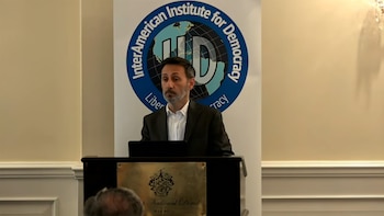 Hombre con barba y traje oscuro en un podio. Detrás, pancarta con el logo del InterAmerican Institute for Democracy. Placa de Trump National Doral Miami