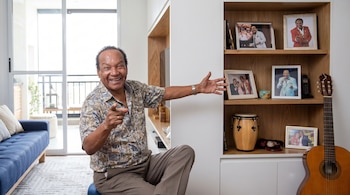 Melcochita sonriente sentado en su sala, señalando hacia adelante, con un sofá azul a la izquierda y un estante con fotos, un bongó y una guitarra a la derecha.