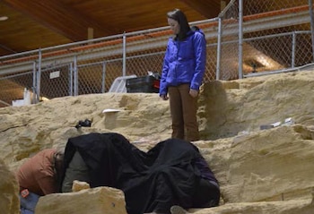 Una mujer con chaqueta azul observa a dos personas agachadas junto a una manta negra sobre una formación de piedra