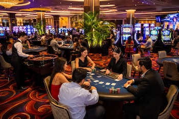 Personas sentadas alrededor de una mesa de cartas con fichas junto a un crupier, una ruleta y máquinas tragamonedas.