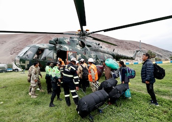 Civiles con equipaje abordan un helicóptero militar sobre hierba, asistidos por policías y militares junto a cerros áridos