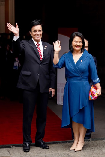 Un hombre con traje y corbata y una mujer con vestido azul y cartera de mano saludan con las manos alzadas en una alfombra roja frente a una puerta