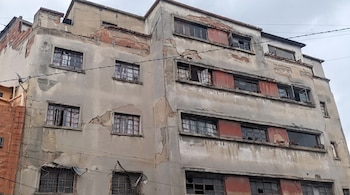 Vista de un edificio de múltiples pisos con fachada de hormigón dañada, yeso desprendido, ladrillos expuestos, ventanas rotas y cables eléctricos cruzados